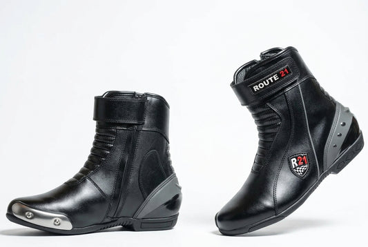 A pair of black leather motorcycle drag racing boots shown from the side and back on a light gray studio background. One boot has a silver metal toe cap, and both display 'ROUTE 21 Apparel' branding.