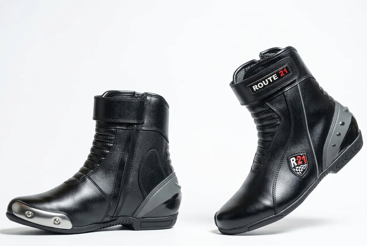 A pair of black leather motorcycle drag racing boots shown from the side and back on a light gray studio background. One boot has a silver metal toe cap, and both display 'ROUTE 21 Apparel' branding.