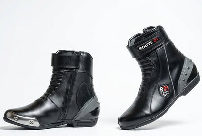 A pair of black leather motorcycle drag racing boots shown from the side and back on a light gray studio background. One boot has a silver metal toe cap, and both display 'ROUTE 21 Apparel' branding.